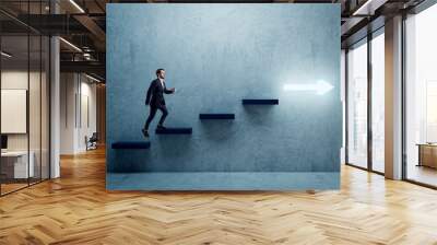 Side view of young business man climbing illuminated arrow stairs to success on concrete wall background in interior. Financial growth, career development and forward concept. Wall mural