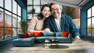 Portrait of Asian mature couple sitting and smiling in living room. wife woman hand hold husband arm from behind and look at camera with happiness and cheerful safty amd insurance family concept Wall mural