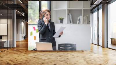 Asian businesswoman talking on mobile phone working on laptop in modern office. Happy business woman talking on mobile phone while analyzing weekly schedule in laptop Wall mural