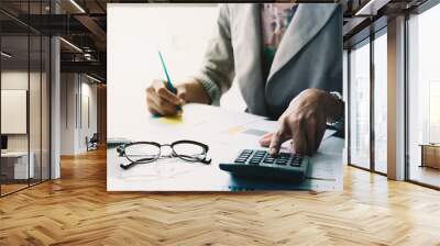 Close up of businessman or accountant hand holding pen working on calculator to calculate business data, accountancy document and laptop computer at office, business concept Wall mural