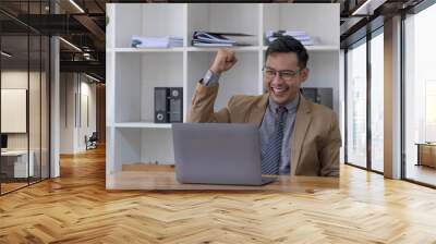 Happy young businessman in suit looking at laptop excited by good news online, lucky successful winner man standing at office desk raising hand in yes gesture celebrating business success win result Wall mural