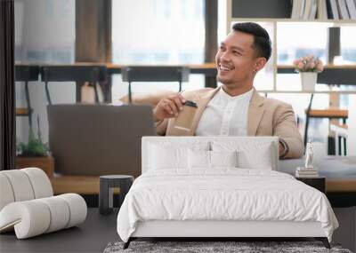 Young Asian businessman in suit and holding hot coffee cup and looking on laptop computer in office. Man in suit using laptop in well-lit workplace. Wall mural