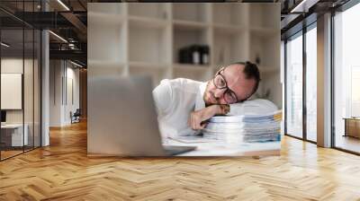 Young unhappy man office worker feeling bored at work, looking at laptop with demotivated face expression while sitting at workplace in office, distracted male worker feeling tired of monotonous job Wall mural