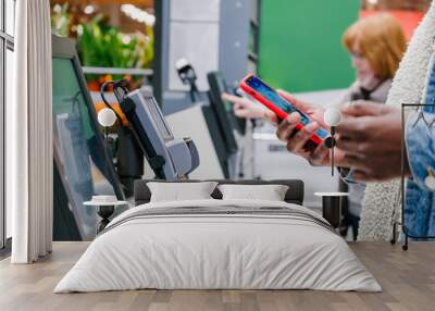 Black male person in warm denim jacket uses smartphone to pay for purchase at self-checkout point in supermarket close view Wall mural