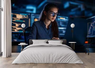 Serious young woman programmer working on laptop in a tech office with multiple monitors displaying code Wall mural