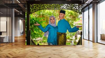 A portrait of young couple of malay muslim in traditional costume during Aidilfitri celebration showing welcome gesture by traditional wooden house Wall mural