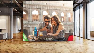 Happy young couple tourists using smartphone sitting at bar restaurant in front of colosseum in rome at sunset with coffee shopping bags smiling having fun texting browsing and sharing pictures Wall mural