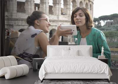 Two happy young woman tourists sitting at the table of a bar restaurant in front of the Colosseum in Rome using smartphone and laptop computer while having a glass of italian red wine. Stylish Wall mural