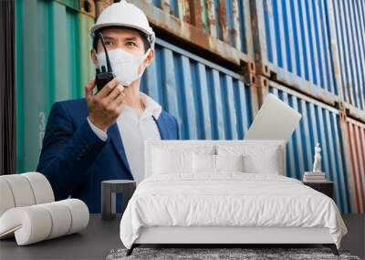 Logistic engineer manager man wearing protective mask holding laptop and talking with walkie talkie working at container warehouse construction site.  Wall mural