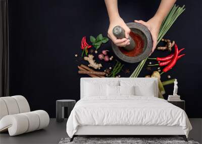 The Art of Thai Cuisine - Thai lady’s hands hold stone granite pestle with mortar and red curry paste ingredient together with fresh herbs and spices on classic dark background at top view angle. Wall mural