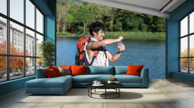 Thirsty hiker with backpack opening bottle and drinking water by scenic lake. Young Asian man. Wall mural