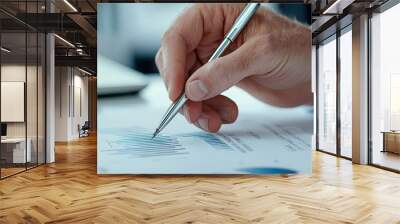 A close-up of a hand holding a pen, analyzing financial graphs on paper, illustrating data analysis and strategy in a professional setting. Wall mural