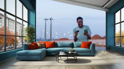 happy african youth using his phone and credit card to make payment online. Buying and shopping concept Wall mural