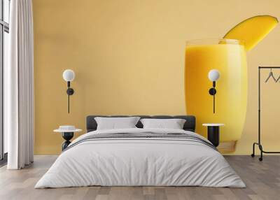 A tall glass of mango smoothie with a slice of mango on the rim, sitting on a yellow background. Wall mural