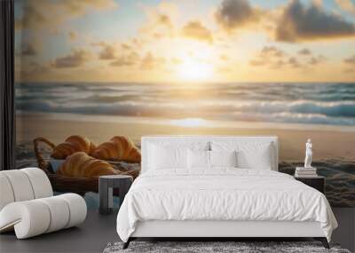 A wicker basket filled with croissants, a cup of coffee, and a pastry sits on a white cloth beside the ocean at sunrise. Wall mural