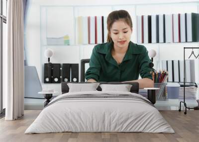 A cheerful, confident Asian businesswoman sits at a reception desk, smiling as she talks on her smartphone. Surrounded by modern technology, she manages client communications and multitasks between he Wall mural