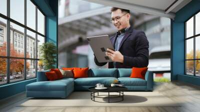 A cheerful young Asian businesswoman in a modern city, smiling confidently while using her smartphone outdoors, dressed in a professional suit, embodying success and digital connectivity. Wall mural