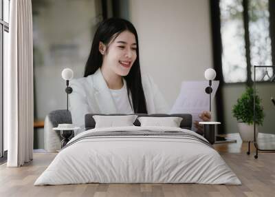 A happy Asian female businessman is at a modern desk. Financial reports and online documents using laptops She analyzed the organization's strategy and cooperated with her team for success. Wall mural