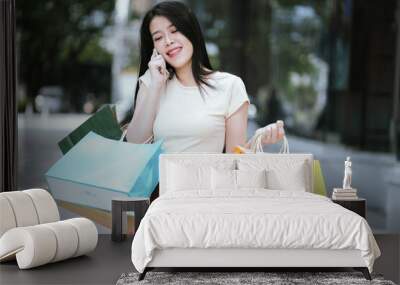 A modern young woman smiles as she walks in front of a shopping mall, each carrying shopping bags. One is checking her smartphone, enjoying the modern city life and shopping experience. Wall mural