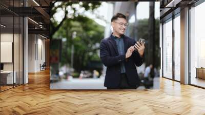 Confident Asian businessman in modern city, smiling while using smartphone outdoors, wearing professional suit, representing success and connectivity in digital age. Wall mural