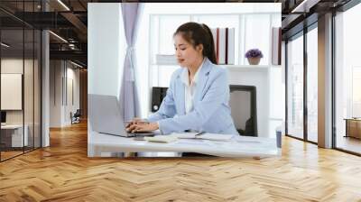 Confident Asian businesswoman in modern office smiling while working on laptop, analyzing reports, planning marketing strategies and leading with energy and positivity. Wall mural