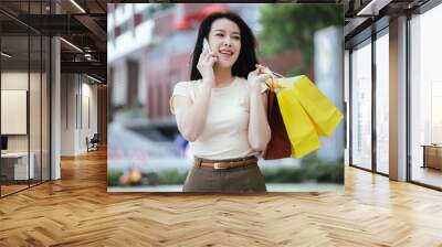 Happy Asian woman sitting on outdoor bench, sipping juice and holding shopping bags. She is relaxing, surfing the web on her smartphone, enjoying a relaxing weekend in the city after a day of shopping Wall mural