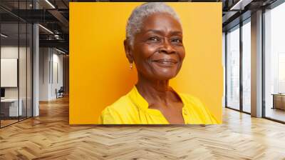 a Black elderly person in a yellow blouse, looking content with a gentle smile on a yellow studio background Wall mural