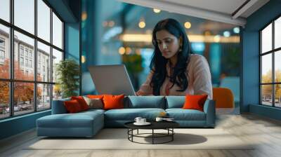 An Indian businesswoman concentrates on her work, typing on a laptop in a sleek office space, illustrating diligence and commitment. Wall mural