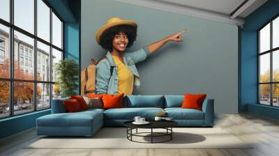 Happy female tourist points at copy space background. Beautiful African American woman with travel bag in casual clothes standing by gray wall, pointing to side and smiling. Holiday trip concept Wall mural