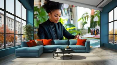 Young adult African American female fashion designer at her desk, examining fabric swatches, with a modern studio filled with design tools and a serene green background. Wall mural