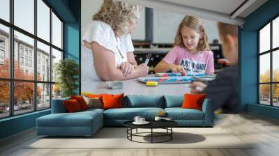 A girl in a pink shirt is sitting at a table with a woman and a boy. The girl is learning with blocks and the woman is watching her. Scene is playful and lighthearted Wall mural