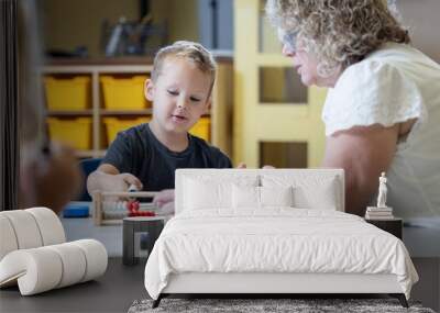 A young boy is sitting at a table with a teacher, who is helping him with a math problem. The woman is wearing glasses and has a serious expression on her face. The boy is focused on the task at hand Wall mural