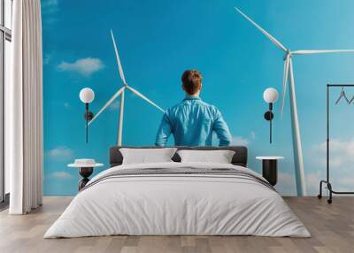 A man stands in front of two wind turbines, looking up at them Wall mural