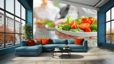 A woman is holding a plate of salad with broccoli, tomatoes, and lettuce Wall mural