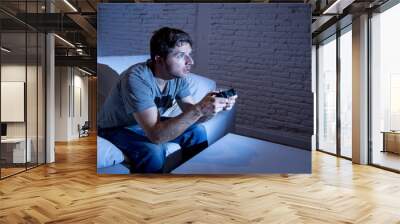 young excited man at home sitting on living room sofa playing video games using remote control joystick Wall mural