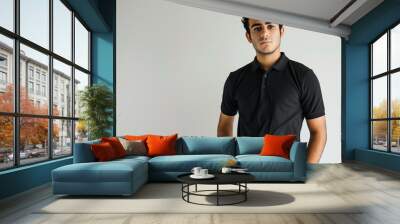 young Middle Eastern man standing with his hands in his pockets, wearing a black polo shirt and black pants, short curly dark hair, medium skin tone, plain white background Wall mural