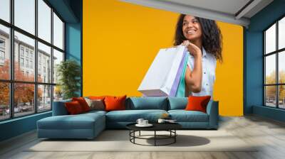 Attractive african american young woman with perfect smile is posing with a pile of paper shopping bags in hand and looking aside, isolated on yellow background Wall mural