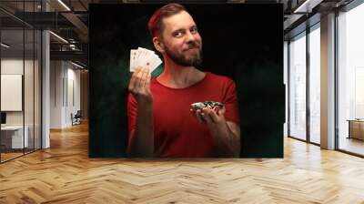 Studio portrait of smiling handsome young bearded man posing with a fan of playing cards and pile of poker chips over black background with smoke effect Wall mural