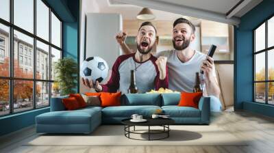 Two happy buddies sitting on sofa, watching football play live broadcast and celebrating their favourite team victory Wall mural