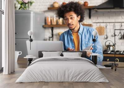 Young confident African American man working from home sitting in the kitchen with his laptop computer, participating in video conference, having online meetup with his coworkers, employee or client Wall mural