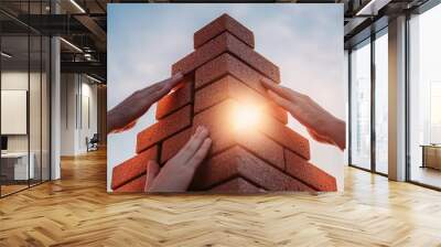 Two hands are positioned to build a brick structure, with a glowing light emerging from the top, symbolizing teamwork and progress against a clear sky. Wall mural