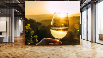 Captivating hand holding white wine glass against lush, rolling vineyards bathed in warm sunset glow, an elegant and scenic display of relaxation. Wall mural