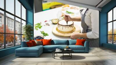 A confectioner squeezes liquid chocolate from a pastry bag onto a white cream biscuit cake on a wooden stand. The concept of homemade pastry, cooking cakes. Wall mural