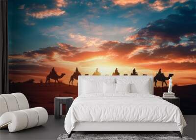 A group of camels are walking across a desert at sunset Wall mural