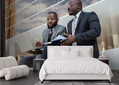 Business meeting of friends outdoors. Two dark-skinned men in suits are sitting on a bench near a city building with a laptop and talking. Partnership and corporate relations Wall mural