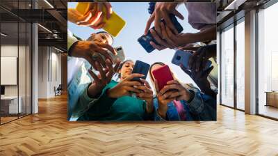 Diverse group of young people using mobile phone device standing together in circle outdoors. Millennial friends addicted to social media app, betting or playing video game on platform online. Wall mural