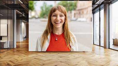 Happy young caucasian girl smiling at camera standing at city street. Outside portrait of joyful beautiful italian woman over urban background. Wall mural