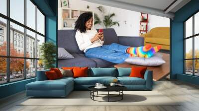 Millennial african american woman using mobile phone sitting on couch at home - Mixed race brazilian female chatting on smartphone Wall mural