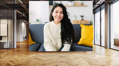 Portrait of cheerful young latin american woman smiling at camera sitting on sofa at home. Wall mural