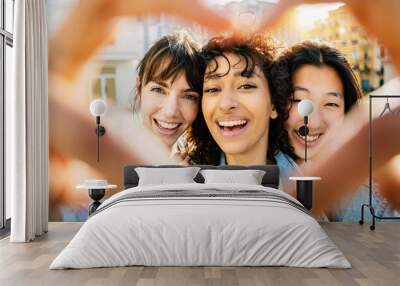 Three happy diverse young women drawing heart shape with hands outdoors. Smiling portrait of multi ethnic female friends having fun at city street. Love, youth lifestyle and friendship concept Wall mural